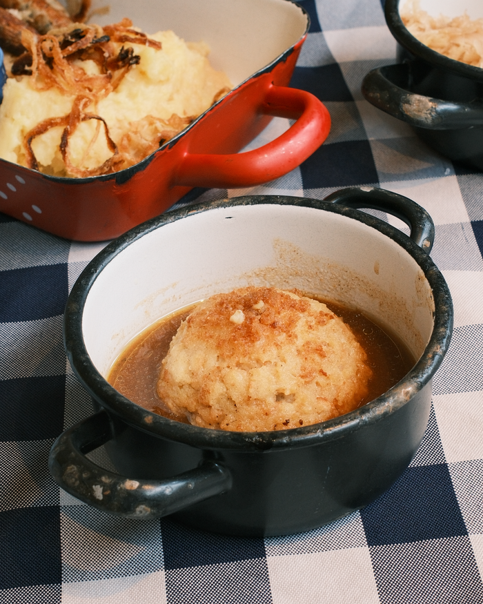 Warm Bavarian bread dumpling served in broth with mashed potatoes on checkered tablecloth.